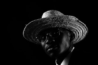 A person wearing a textured straw hat and glasses, captured in dramatic low-key lighting against a dark background. The light highlights the woven pattern of the hat and softly illuminates part of the face, creating strong contrasts.