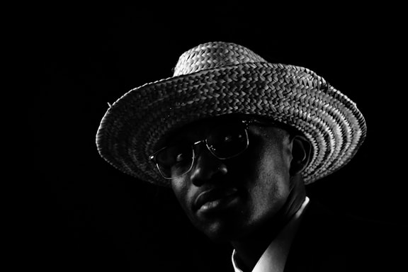 A person wearing a textured straw hat and glasses, captured in dramatic low-key lighting against a dark background. The light highlights the woven pattern of the hat and softly illuminates part of the face, creating strong contrasts.
