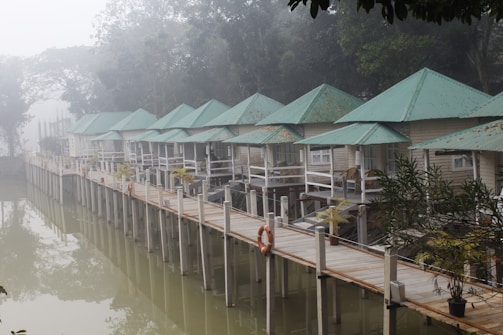 A serene lakeside scene with several small, quaint cabins built on stilts over the water, featuring green metal roofs and light-colored walls. The cabins are connected by a wooden walkway with white railings, lined with potted plants. A life preserver hangs on the railing, and the entire area is enveloped in a misty, foggy atmosphere, with trees visible in the background.