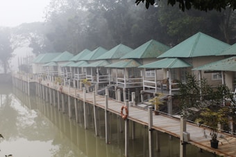 A serene lakeside scene with several small, quaint cabins built on stilts over the water, featuring green metal roofs and light-colored walls. The cabins are connected by a wooden walkway with white railings, lined with potted plants. A life preserver hangs on the railing, and the entire area is enveloped in a misty, foggy atmosphere, with trees visible in the background.
