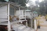 A row of elevated wooden cabins with green-tiled roofs stands alongside a wooden walkway. Each cabin has an open front porch furnished with a couple of wicker chairs and a small table. The cabins are built on stilts over water, and there are small potted plants along the walkway. Dense trees form the background.