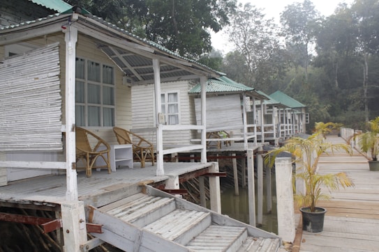 A row of elevated wooden cabins with green-tiled roofs stands alongside a wooden walkway. Each cabin has an open front porch furnished with a couple of wicker chairs and a small table. The cabins are built on stilts over water, and there are small potted plants along the walkway. Dense trees form the background.