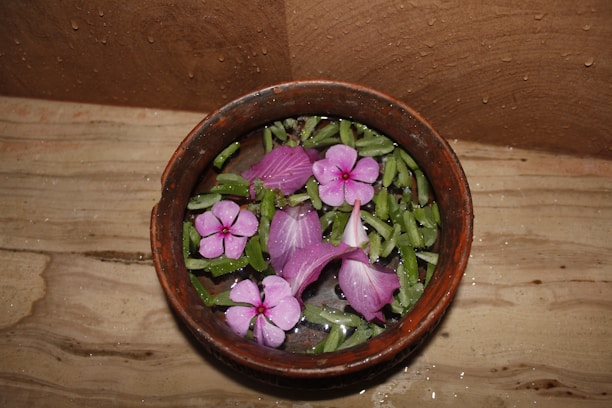 A rustic clay pot filled with water contains several floating pink flowers and pieces of green foliage. The background features a wooden surface with visible texture and water droplets, adding a natural and earthy feel.