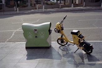A yellow electric bicycle is parked next to a green and white recycling bin on a sidewalk, with a road in the background. The bike has a helmet on the handlebars and appears to be for public rental. The scene is peaceful and suggests a sunny day.
