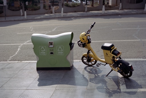 A yellow electric bicycle is parked next to a green and white recycling bin on a sidewalk, with a road in the background. The bike has a helmet on the handlebars and appears to be for public rental. The scene is peaceful and suggests a sunny day.