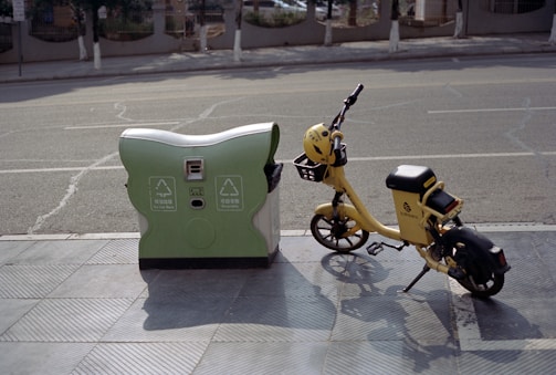 A yellow electric bicycle is parked next to a green and white recycling bin on a sidewalk, with a road in the background. The bike has a helmet on the handlebars and appears to be for public rental. The scene is peaceful and suggests a sunny day.