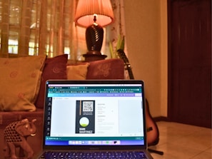 A cozy room with a laptop placed on a couch, displaying a digital design on the screen. The background includes decorative cushions, a warm-lit lamp on a table, and a guitar resting against the wall. An ornate small elephant figurine is also visible next to the laptop.
