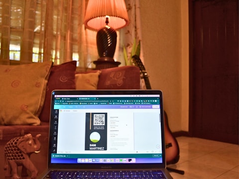 A cozy room with a laptop placed on a couch, displaying a digital design on the screen. The background includes decorative cushions, a warm-lit lamp on a table, and a guitar resting against the wall. An ornate small elephant figurine is also visible next to the laptop.