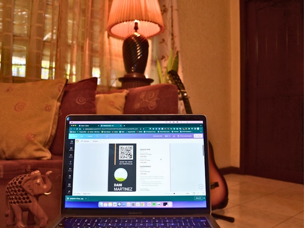 A cozy room with a laptop placed on a couch, displaying a digital design on the screen. The background includes decorative cushions, a warm-lit lamp on a table, and a guitar resting against the wall. An ornate small elephant figurine is also visible next to the laptop.