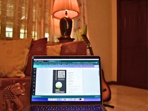 A cozy room with a laptop placed on a couch, displaying a digital design on the screen. The background includes decorative cushions, a warm-lit lamp on a table, and a guitar resting against the wall. An ornate small elephant figurine is also visible next to the laptop.