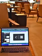 A laptop resting on a wooden table displays a graphic design software with a black card featuring a letter 'C' and a QR code. The background reveals a modern cafe setting with cushioned chairs, wooden tables, and soft lighting, all contributing to a cozy atmosphere.