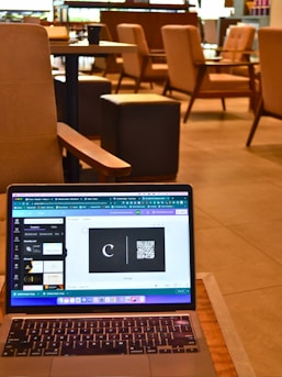 A laptop resting on a wooden table displays a graphic design software with a black card featuring a letter 'C' and a QR code. The background reveals a modern cafe setting with cushioned chairs, wooden tables, and soft lighting, all contributing to a cozy atmosphere.