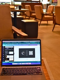 A laptop resting on a wooden table displays a graphic design software with a black card featuring a letter 'C' and a QR code. The background reveals a modern cafe setting with cushioned chairs, wooden tables, and soft lighting, all contributing to a cozy atmosphere.