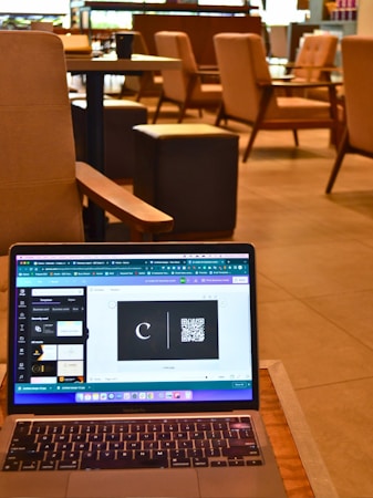 A laptop resting on a wooden table displays a graphic design software with a black card featuring a letter 'C' and a QR code. The background reveals a modern cafe setting with cushioned chairs, wooden tables, and soft lighting, all contributing to a cozy atmosphere.