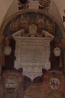 A classical wall monument with intricate carvings and inscriptions sits surrounded by decorative coats of arms. The monument features a large central plaque with Latin text, topped by a sculpted bust and ornate architectural elements. Beneath this, colorful heraldic shields add visual interest, featuring emblematic designs and motifs.