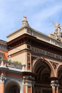 The image depicts part of a historic architectural structure adorned with classical sculptures. An angelic statue with wings is prominently placed on the rooftop, accompanied by another figure looking outward. The building showcases intricate carvings and ornate details, with a color palette of warm tones, including terracotta and beige.