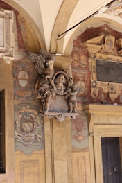 An ornate interior wall featuring a sculpted relief with a central bust framed by angels, possibly cherubs, above an inscribed plaque. The wall is richly decorated with elaborate frescoes, heraldic symbols, and shields, with a color palette dominated by earth tones. Architectural elements like columns and arches add to the historical and artistic ambiance.