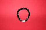 A round bracelet made of black beads arranged on a red background. Some beads are interspersed with metallic, decorative rings, and the bracelet features a yin-yang symbol as part of its closure.