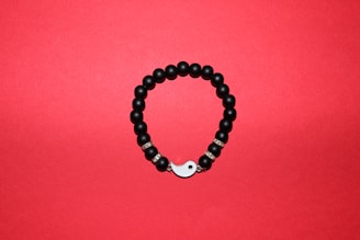 A round bracelet made of black beads arranged on a red background. Some beads are interspersed with metallic, decorative rings, and the bracelet features a yin-yang symbol as part of its closure.