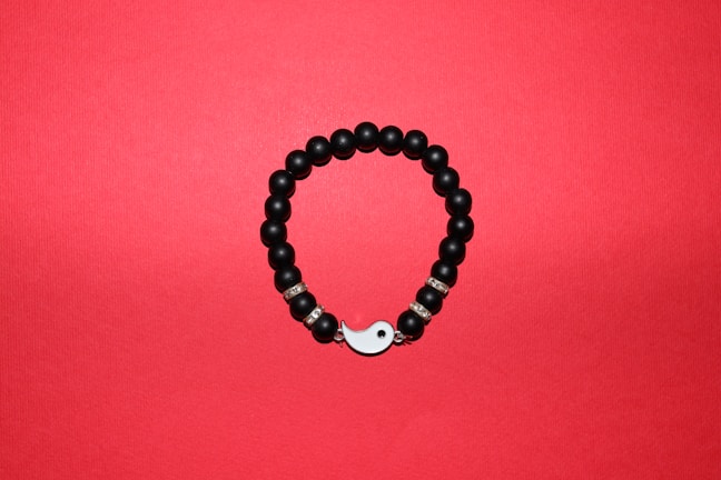 A round bracelet made of black beads arranged on a red background. Some beads are interspersed with metallic, decorative rings, and the bracelet features a yin-yang symbol as part of its closure.