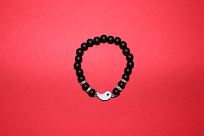 A round bracelet made of black beads arranged on a red background. Some beads are interspersed with metallic, decorative rings, and the bracelet features a yin-yang symbol as part of its closure.