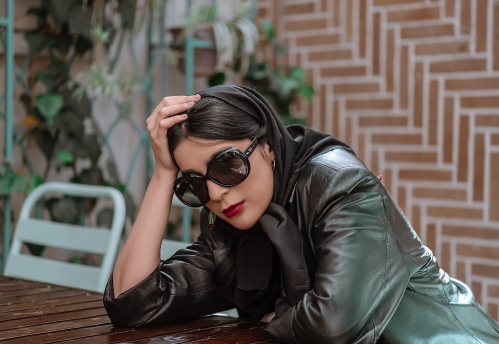 a woman in a leather jacket and sunglasses leaning on a table