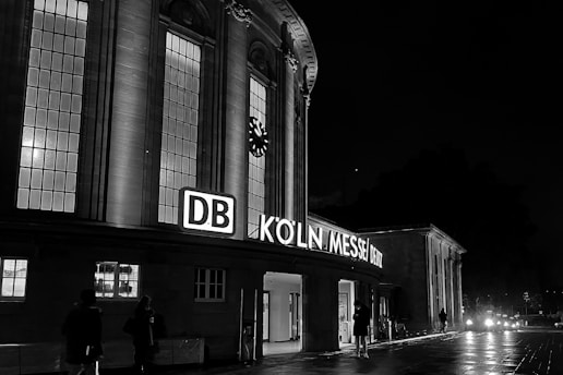 An illuminated building facade at night with large windows and a prominent clock. The building displays neon signage with the text 'DB KÖLN MESSE/DEUTZ'. Several people are walking on the wet pavement, and there is a dark sky above.