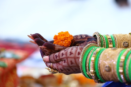 Intricately adorned hands with henna designs hold an orange marigold flower. The wrists are decorated with gold and green bangles. The background is softly blurred, emphasizing the colorful and detailed elements on the hands.
