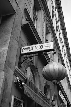 A city street scene featuring a building facade with intricate architectural details. Prominently displayed is a sign reading 'Chinese Food' and a large, traditional Chinese lantern hanging below it. The image is in black and white, emphasizing the textures and lines of the stone building.