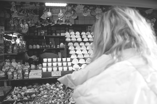 A display of various ceramic items and decorative figures is arranged on shelves in a dimly lit market stall. A person with long hair and a jacket stands in the foreground, facing the stall. The items include cups, bowls, and intricately designed figurines. A price sign is visible, indicating the cost of items in front.