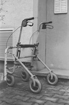 A monochrome image featuring a walker with four wheels parked on a cobblestone pavement next to a building. The walker is positioned leaning against a metal structure for support. The scene includes part of the building wall with a vent and some foliage visible in the background.
