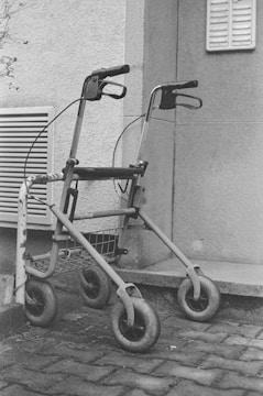 A monochrome image featuring a walker with four wheels parked on a cobblestone pavement next to a building. The walker is positioned leaning against a metal structure for support. The scene includes part of the building wall with a vent and some foliage visible in the background.