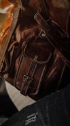 Close-up of a brown leather bag featuring detailed stitching and metallic rivets. The texture of the leather is highlighted by soft lighting.