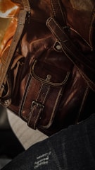 Close-up of a brown leather bag featuring detailed stitching and metallic rivets. The texture of the leather is highlighted by soft lighting.