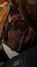 Close-up of a brown leather bag featuring detailed stitching and metallic rivets. The texture of the leather is highlighted by soft lighting.