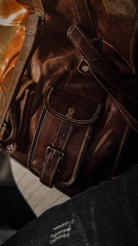 Close-up of a brown leather bag featuring detailed stitching and metallic rivets. The texture of the leather is highlighted by soft lighting.