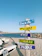 A beach scene featuring a colorful wooden signpost with multiple planks labeled 'Base de Lido', 'Windsurf', 'Optimist', 'Catamaran', and 'Paddle'. The signpost stands on sandy ground near a body of water, with white boats nearby and a clear blue sky overhead.