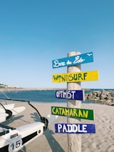 A beach scene featuring a colorful wooden signpost with multiple planks labeled 'Base de Lido', 'Windsurf', 'Optimist', 'Catamaran', and 'Paddle'. The signpost stands on sandy ground near a body of water, with white boats nearby and a clear blue sky overhead.