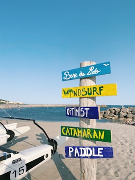 A beach scene featuring a colorful wooden signpost with multiple planks labeled 'Base de Lido', 'Windsurf', 'Optimist', 'Catamaran', and 'Paddle'. The signpost stands on sandy ground near a body of water, with white boats nearby and a clear blue sky overhead.