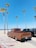 A vintage brown Chevrolet pickup truck is parked next to a modern black pickup on a sandy beach with tall palm trees. The ocean is visible in the background under a clear blue sky.