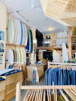 A well-organized surf shop featuring an array of surfboards displayed vertically on the walls, clothing racks with jackets in varying shades, and a wooden counter with a small skateboard leaning against it. The shop's interior includes a mix of wooden and brick elements, with a mezzanine level visible in the background.