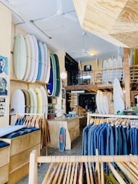 A well-organized surf shop featuring an array of surfboards displayed vertically on the walls, clothing racks with jackets in varying shades, and a wooden counter with a small skateboard leaning against it. The shop's interior includes a mix of wooden and brick elements, with a mezzanine level visible in the background.