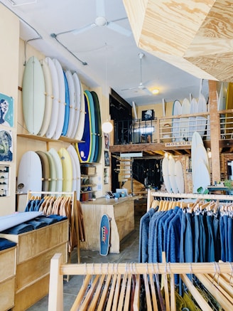 A well-organized surf shop featuring an array of surfboards displayed vertically on the walls, clothing racks with jackets in varying shades, and a wooden counter with a small skateboard leaning against it. The shop's interior includes a mix of wooden and brick elements, with a mezzanine level visible in the background.