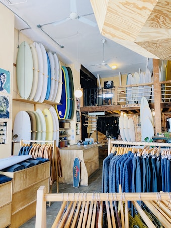 A well-organized surf shop featuring an array of surfboards displayed vertically on the walls, clothing racks with jackets in varying shades, and a wooden counter with a small skateboard leaning against it. The shop's interior includes a mix of wooden and brick elements, with a mezzanine level visible in the background.