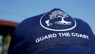 A navy blue cap with a white logo of a tree and a person digging, along with the text 'GUARD THE COAST'. The background is a clear blue sky.