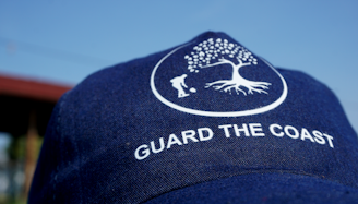 A navy blue cap with a white logo of a tree and a person digging, along with the text 'GUARD THE COAST'. The background is a clear blue sky.