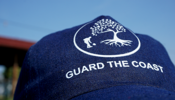 A navy blue cap with a white logo of a tree and a person digging, along with the text 'GUARD THE COAST'. The background is a clear blue sky.
