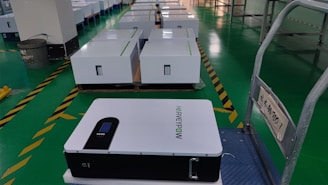 A row of white rectangular boxes, possibly battery or equipment units, are placed on a green tiled floor in a warehouse or manufacturing facility. One unit is prominently visible in the foreground, labeled with the text 'HARVEYPOW'. The area includes safety markings and signage with some text in another language. A trash can is visible in the background.