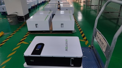 A row of white rectangular boxes, possibly battery or equipment units, are placed on a green tiled floor in a warehouse or manufacturing facility. One unit is prominently visible in the foreground, labeled with the text 'HARVEYPOW'. The area includes safety markings and signage with some text in another language. A trash can is visible in the background.