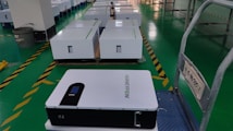 A row of white rectangular boxes, possibly battery or equipment units, are placed on a green tiled floor in a warehouse or manufacturing facility. One unit is prominently visible in the foreground, labeled with the text 'HARVEYPOW'. The area includes safety markings and signage with some text in another language. A trash can is visible in the background.